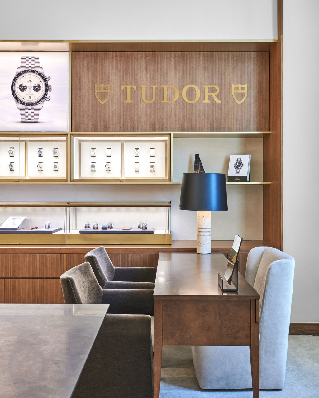 A luxury watch boutique interior featuring a wooden display wall with the gold TUDOR logo, watch cases, and a wooden desk with gray upholstered chairs.
